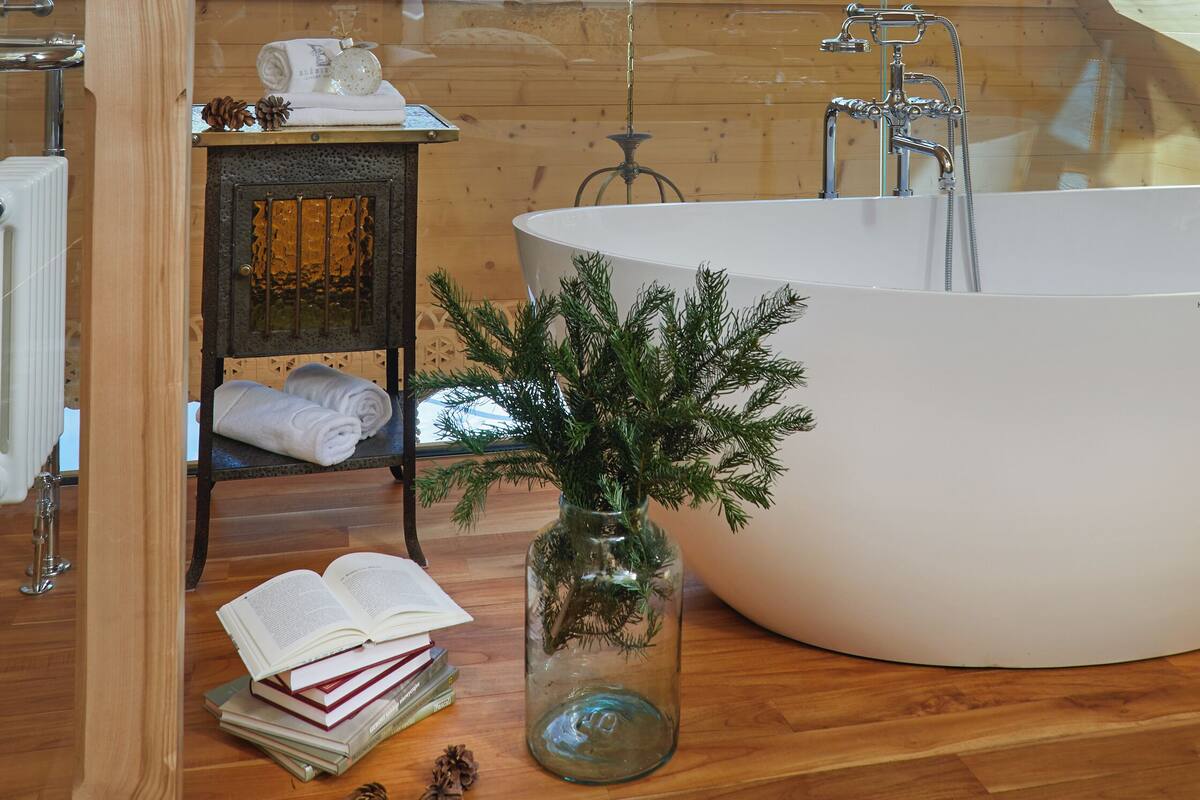 Un interior de madera con tragaluces, una bañera, una silla de madera y una pequeña mesa con libros y piñas.