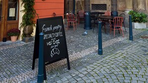 Een straat met kinderafsluiting, terrasjes, een bord met Franse patisserie, speciale koffie en warme chocolademelk, en een gebouw met een rode gevel en een deur met beplanting.