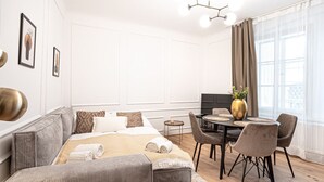 A modern living room with a sofa, a dining table with chairs, a small side table, and a large window with curtains.