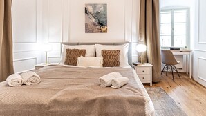 A neatly made bed with a beige blanket and pillows, a desk with a chair, and a window with curtains.