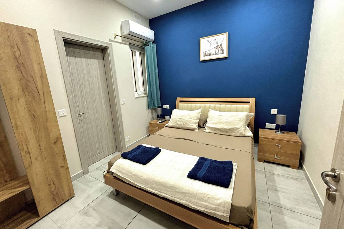 A bedroom with a bed, bedside tables, a nightstand, a window with curtains, an air conditioning unit, and a wall-mounted picture.