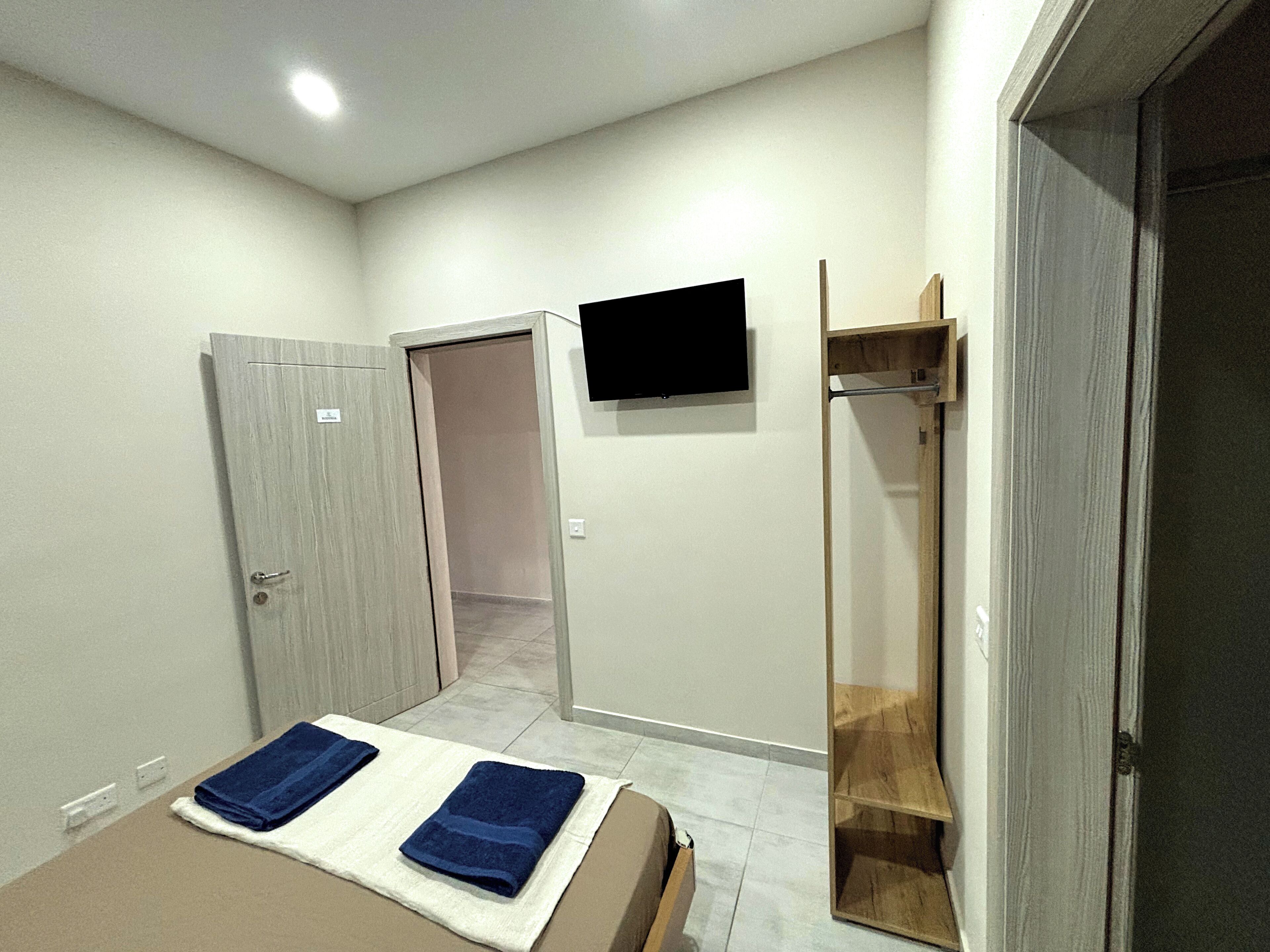 Un dormitorio con cama, dos toallas, televisor, estante de madera y puerta que da a otra habitación.