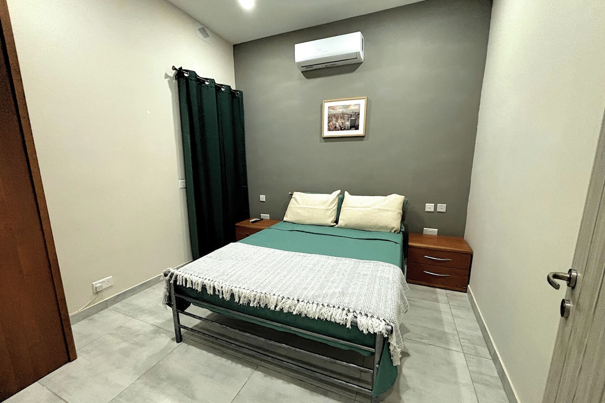 A bedroom with a bed, a nightstand, a drawer, a curtain, an air conditioner, and a framed picture on the wall.