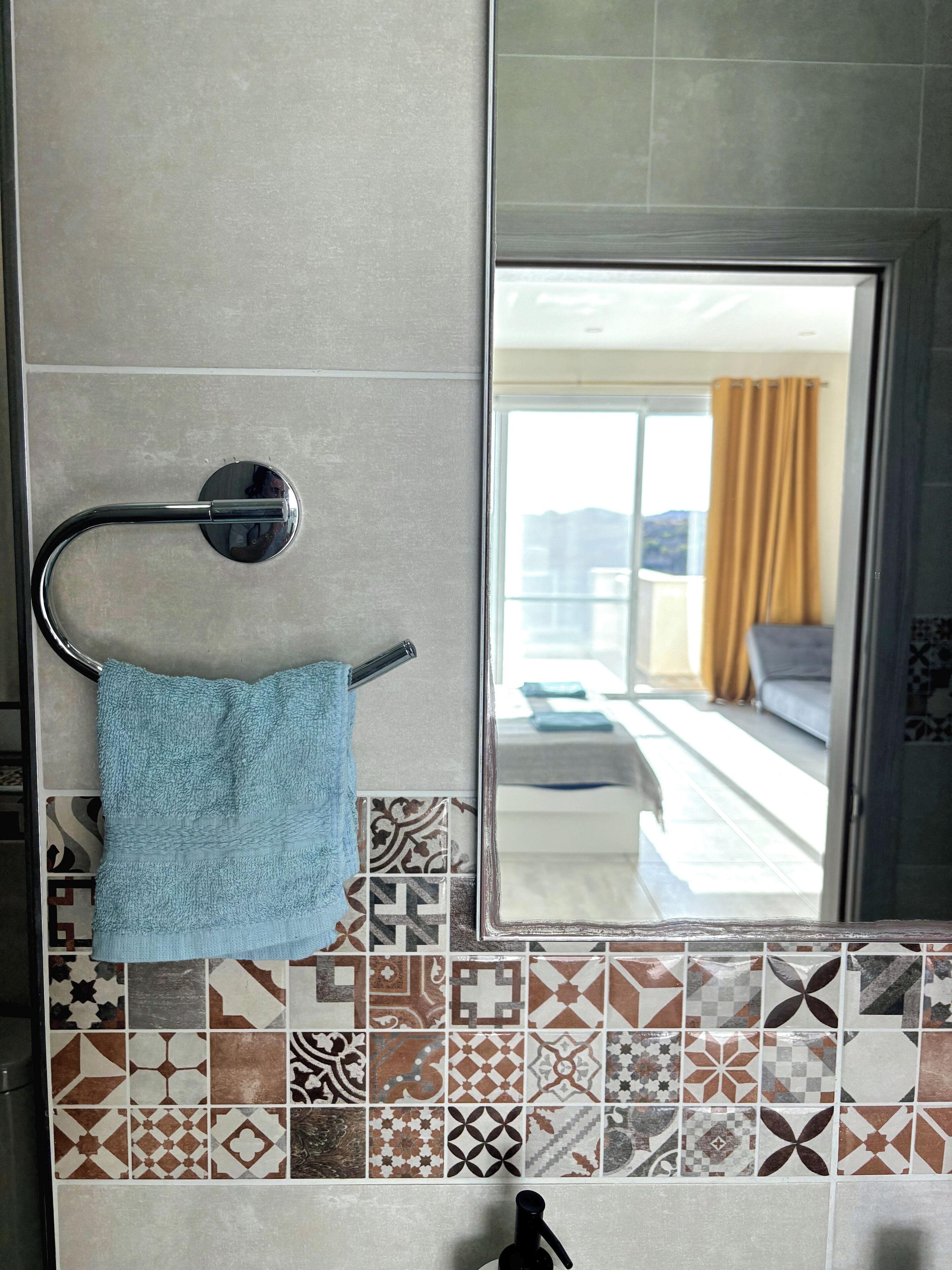 A bathroom with a towel rack, patterned tiles, and a large mirror reflecting a room with a sofa and curtains.