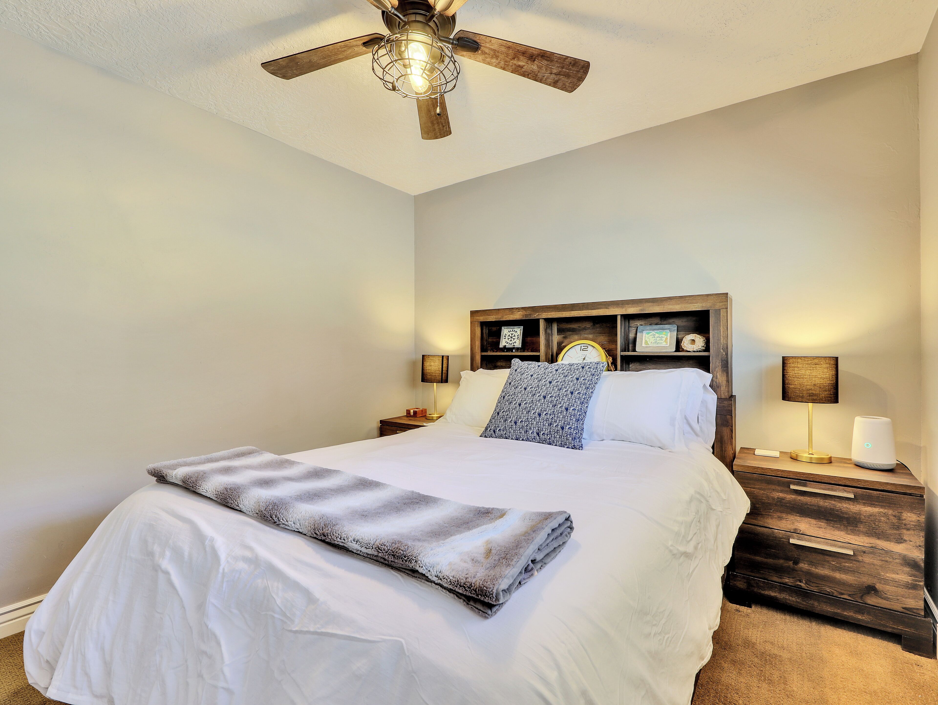 A neatly made bed with a gray blanket, wooden headboard, bedside lamps, and a ceiling fan.