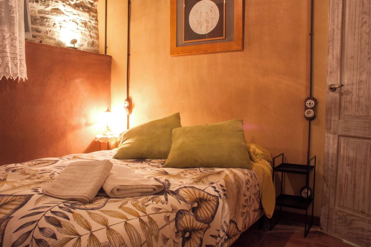 A bedroom with a floral bedspread, two pillows, a bedside lamp, a framed picture on the wall, and a door with a light switch.