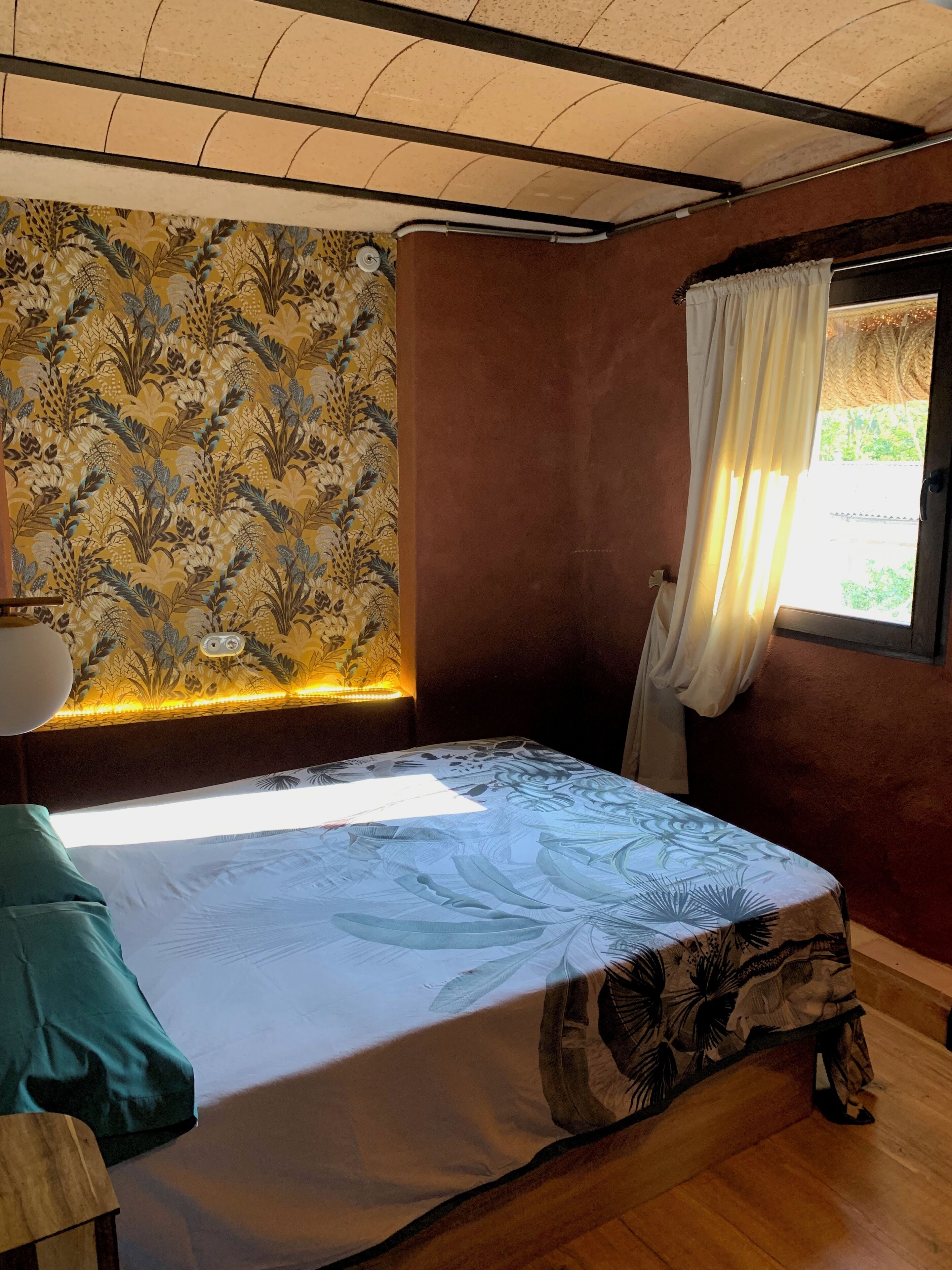 Una habitación con cama, ventana con cortinas, pared con estampado floral y techo con vigas a la vista.