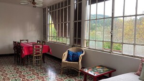 A room with a dining table and chairs, a wicker chair, a small table with a board game, and large windows with a view of greenery. - Mas Cévenol à Proximité du Village et de la Rivière (Lasalle)