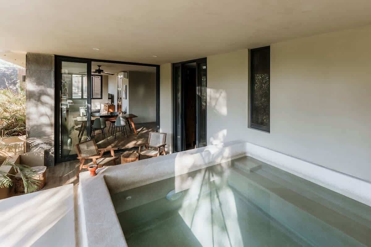 A modern indoor pool area with a glass enclosure, a dining area with chairs and a table, and a large window.