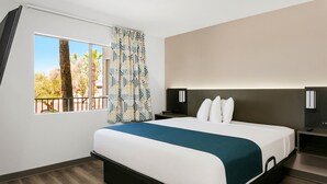 A modern hotel room with a large bed, a nightstand, a TV, and a window with a view of trees and a balcony. - Studio 6 Tempe, AZ – Elliot Road (Tempe)