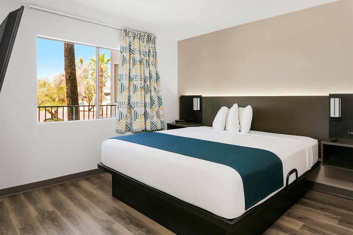 A modern hotel room with a large bed, a nightstand, a TV, and a window with a view of trees and a balcony.
