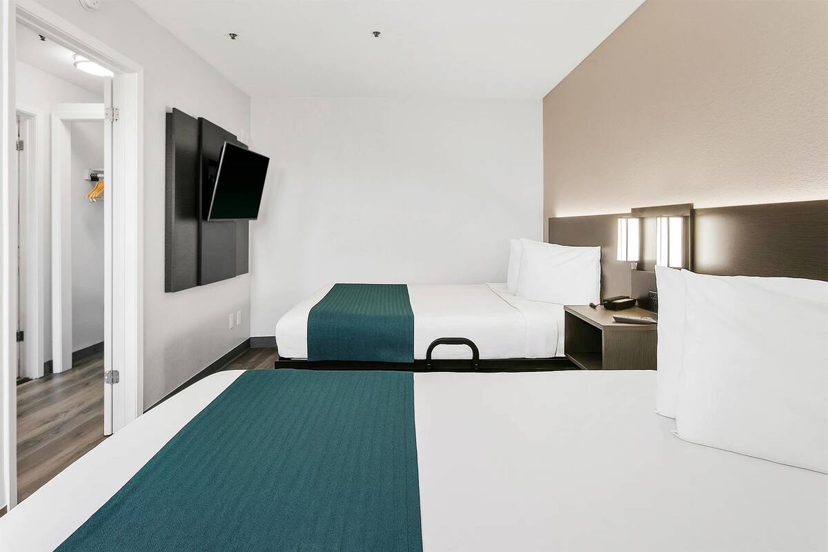 A hotel room with two beds, a flat-screen TV mounted on the wall, a bedside table with a lamp, and a door leading to a hallway.