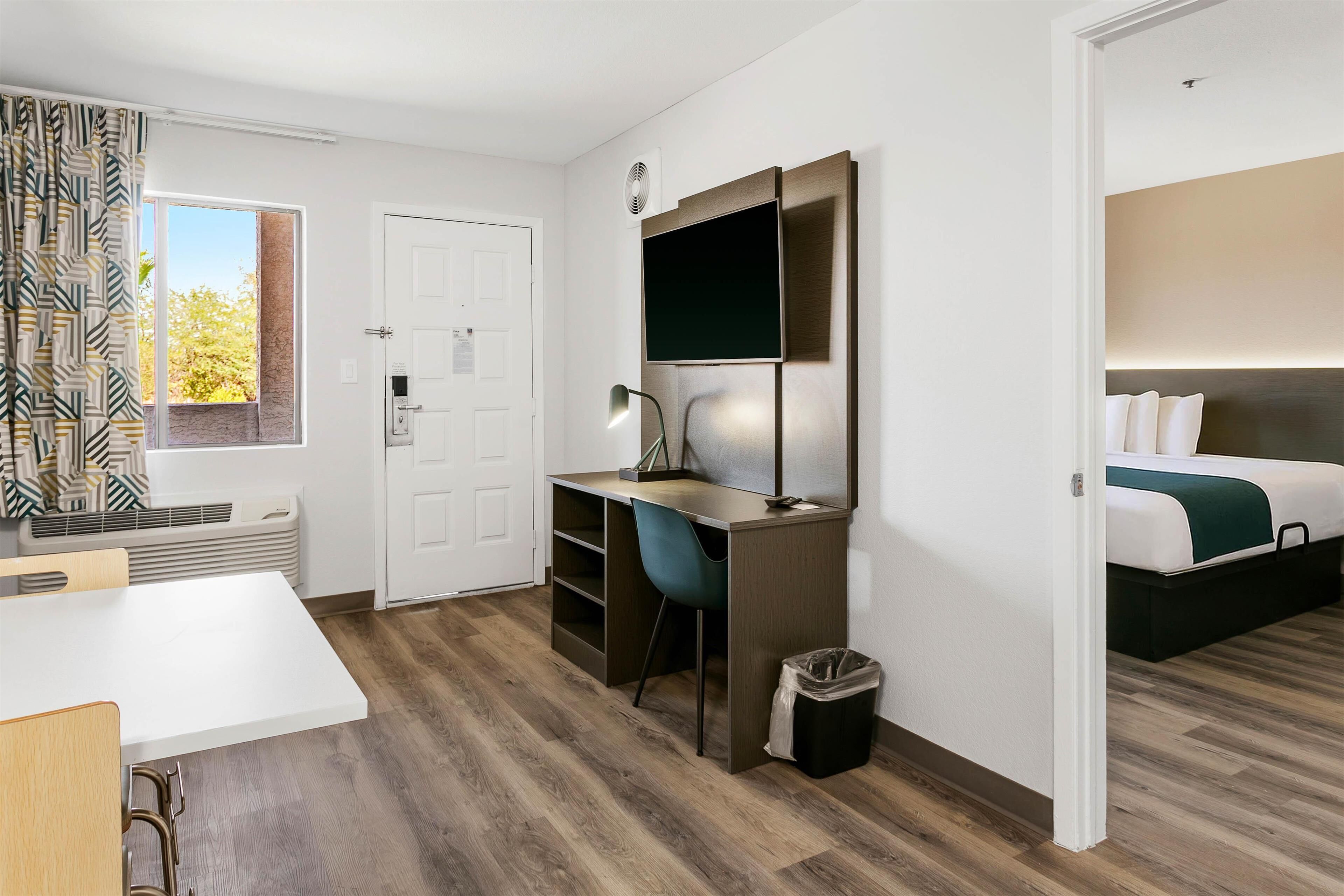 Una habitación de hotel moderna con piso de madera, un escritorio con silla, una cama con un plaid verde y una ventana con cortinas estampadas.