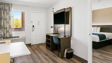 A modern hotel room with a wooden floor, a desk with a chair, a bed with a green throw, and a window with patterned curtains.