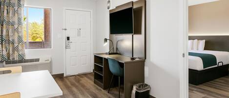 Una habitación de hotel moderna con piso de madera, un escritorio con silla, una cama con un plaid verde y una ventana con cortinas estampadas.