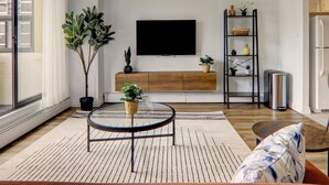 A modern living room with a large TV, wooden cabinet, and a tall plant shelf. - Stylish DT Views at Stampede w/ Free Parking, AC (Calgary)