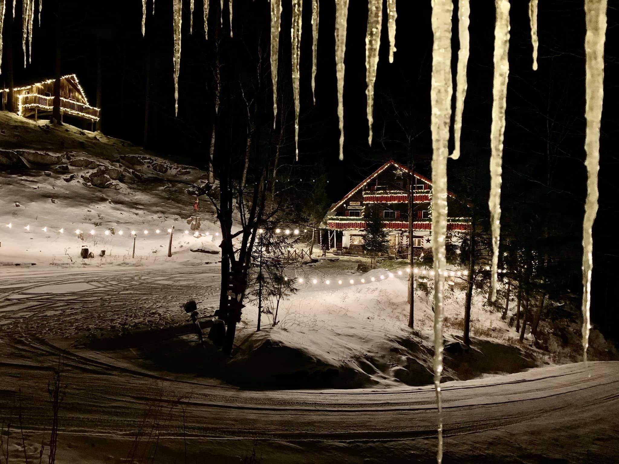 Un paysage enneigé, avec une cabane illuminée par des guirlandes lumineuses, des glaçons pendants des arbres et une motoneige garée à proximité.