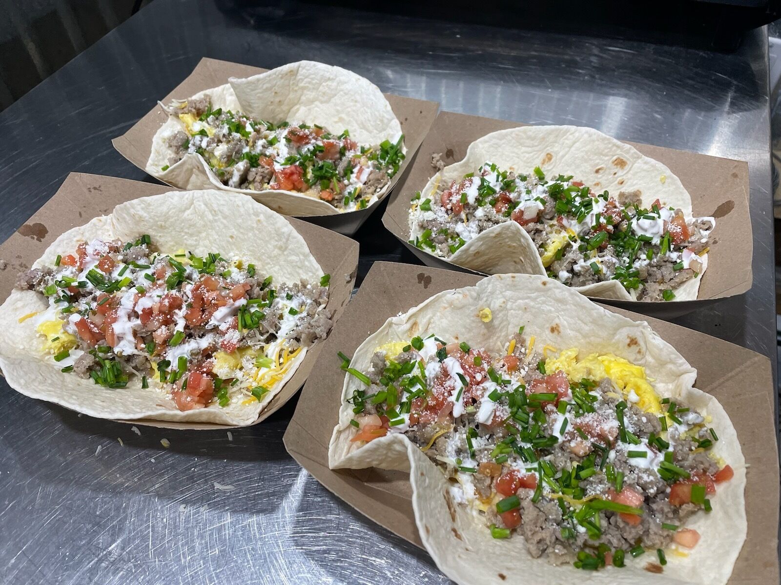 Four tacos with various toppings on a metal surface.