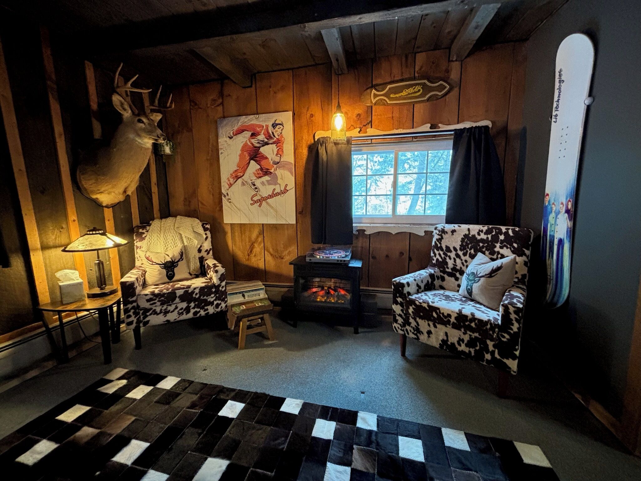 A cozy cabin interior with a fireplace, two animal-themed chairs, a mounted deer head, and a poster of a skier.