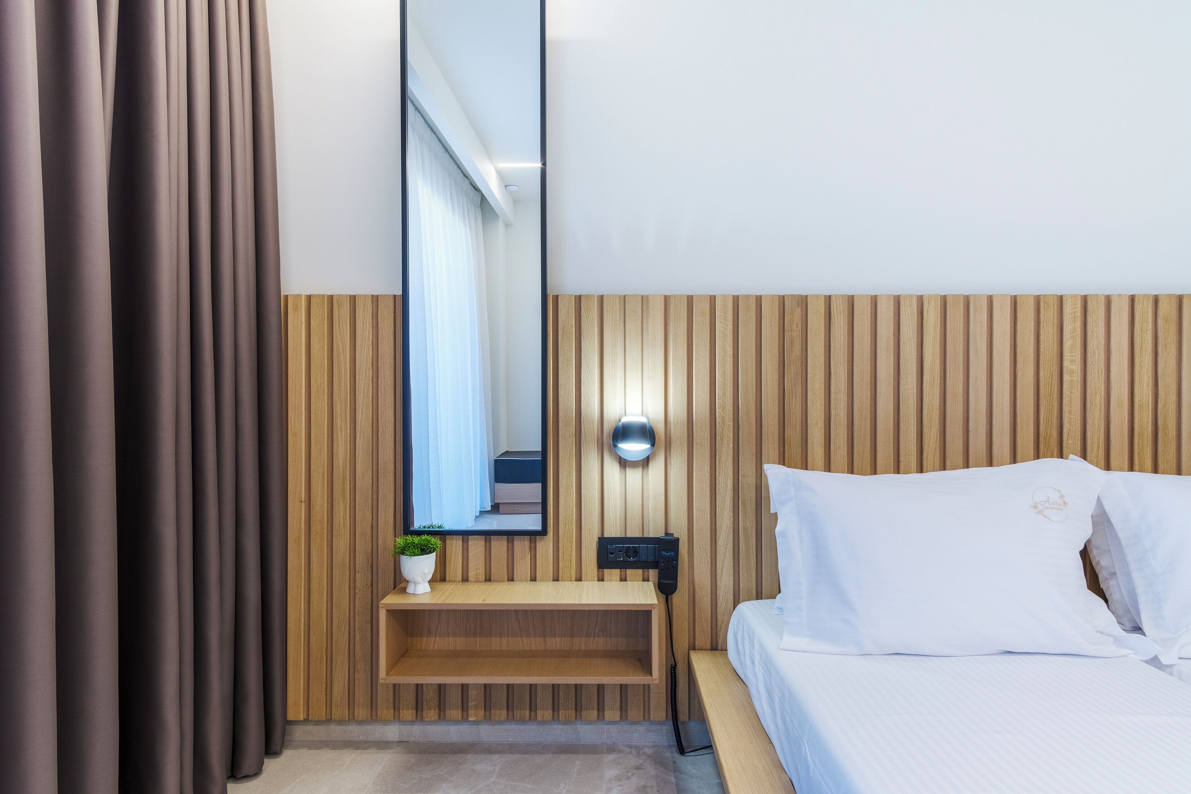 Una habitación de hotel moderna con cabecera de madera, una mesita de noche con una planta, un espejo grande y una cama blanca.