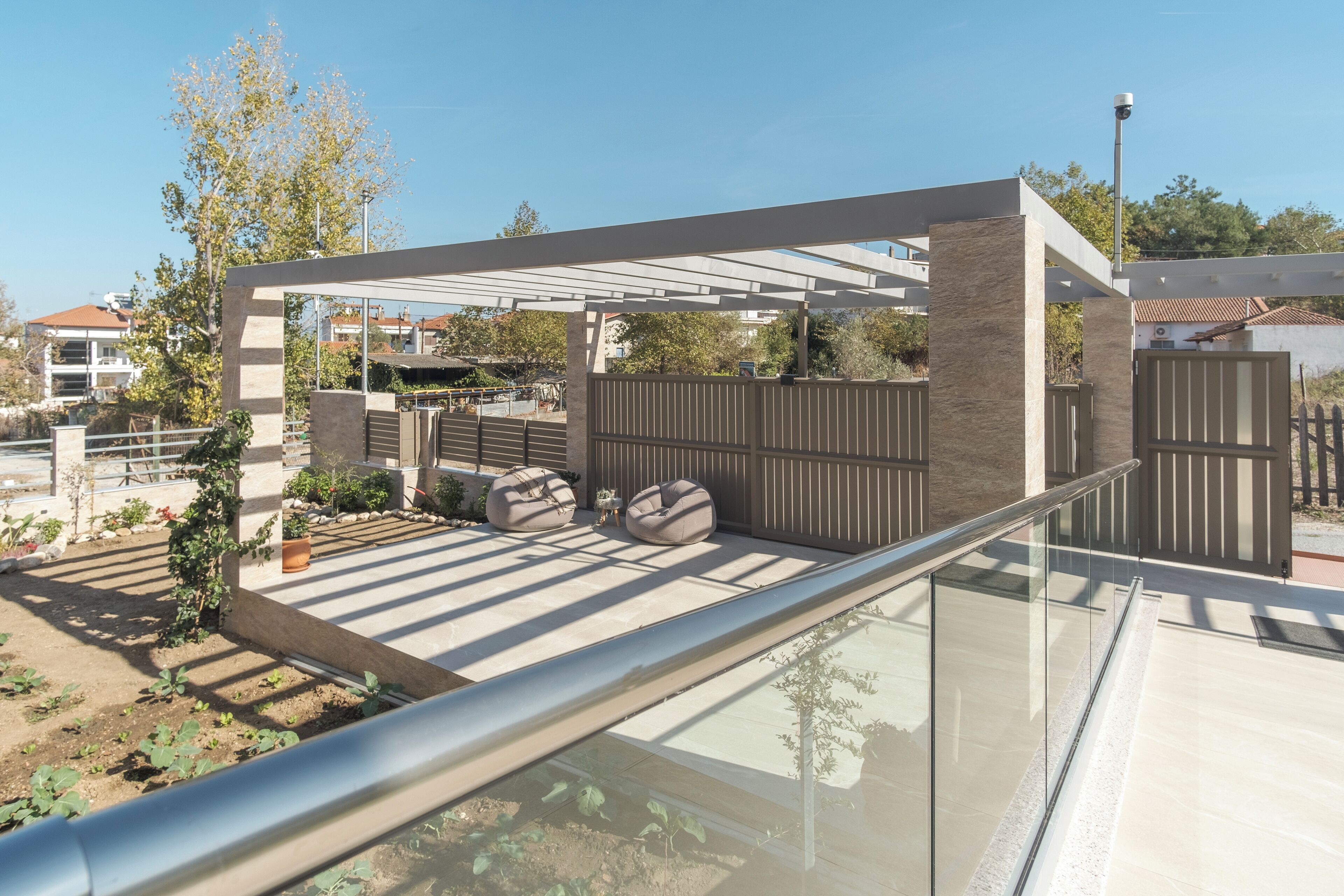 A modern outdoor patio with a glass railing, a seating area with cushions, and a garden with plants and a small tree.
