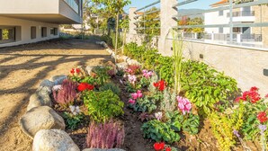 A landscaped garden with various plants and flowers, a dirt path, and a building with balconies in the background. - Aura Luxury Suites - Olympiada Cozy Getaways (Aristotelis)
