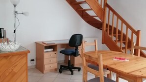 Una habitación con escritorio de madera, silla y mesa de comedor. Se ve una escalera al fondo.