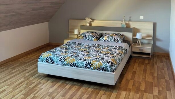 Un dormitorio con techo inclinado, una cama con colcha estampada, una mesita de noche con lámpara y un aplique.
