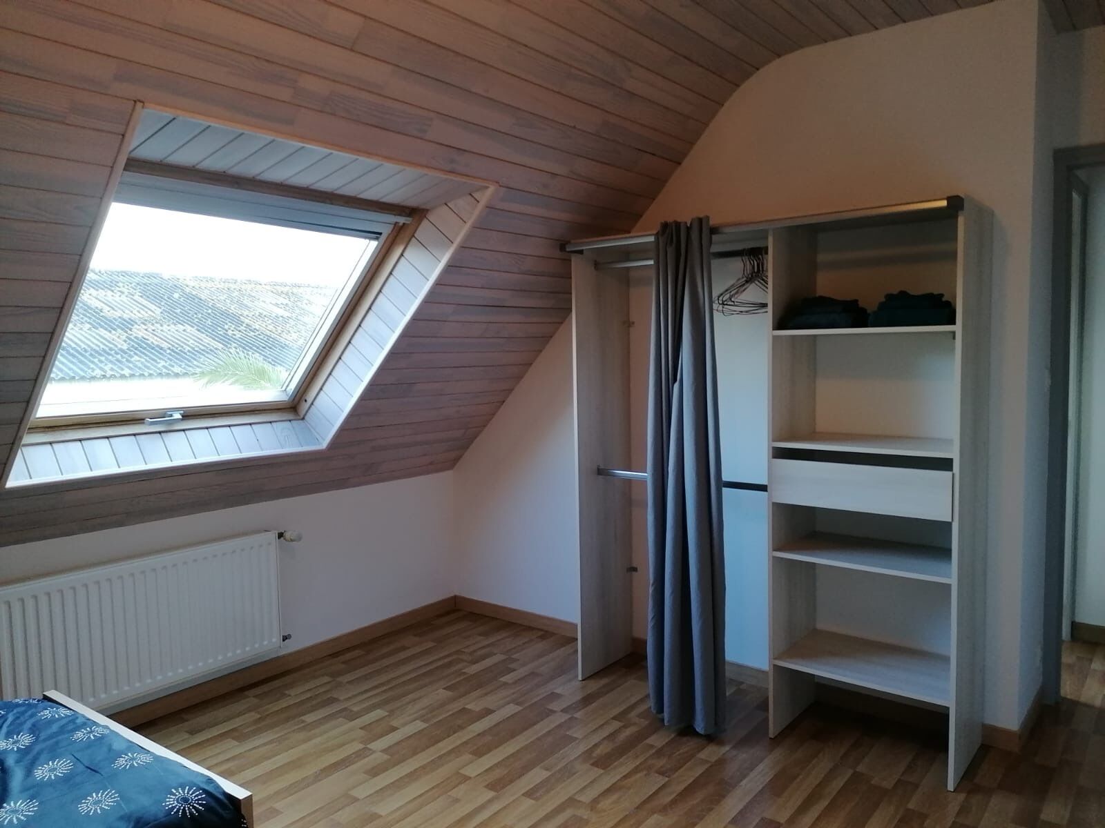 Una habitación con techo inclinado, tragaluz, radiador, cama con cubrecolcha estampada y armario con estantes.