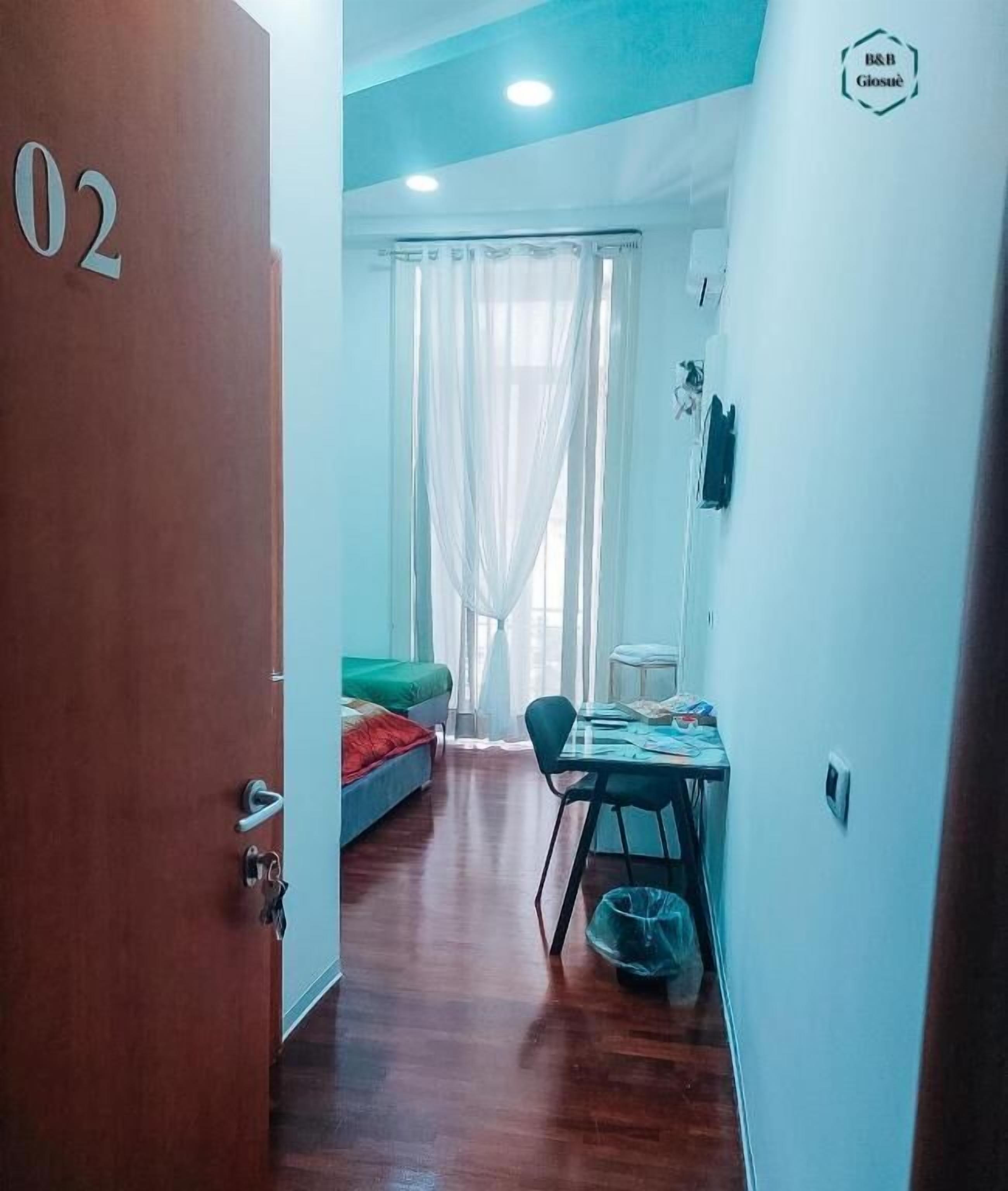 A room with a wooden floor, a bed with a green cover, a table with papers, and a window with sheer curtains.