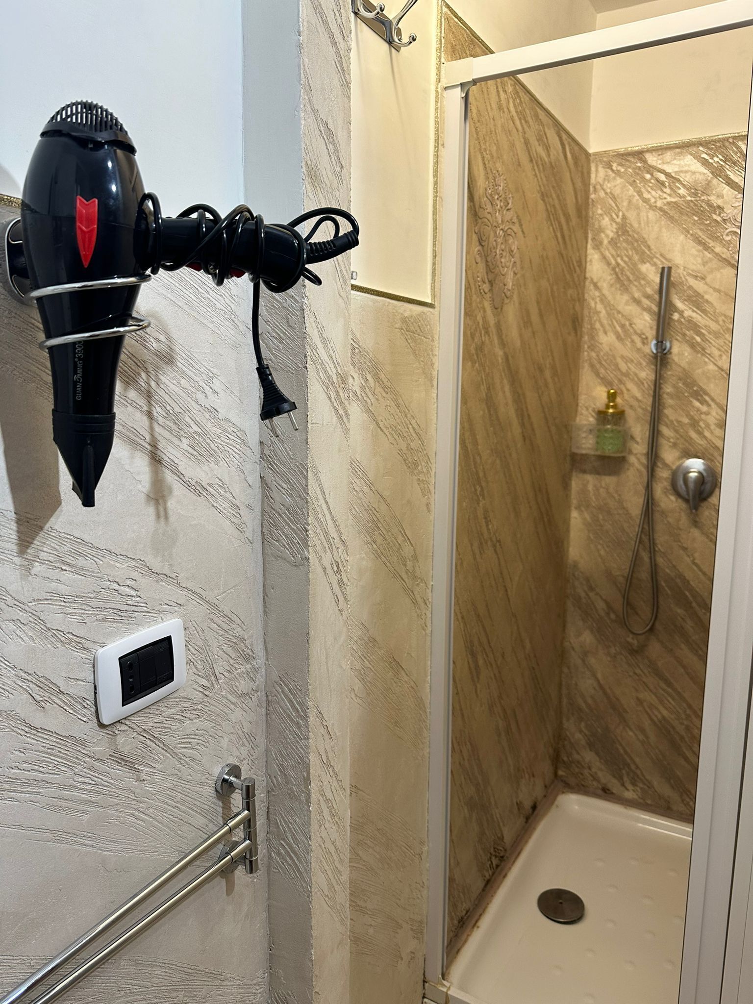 A bathroom with a hairdryer mounted on the wall, a shower with a glass door, and a small shelf with toiletries.
