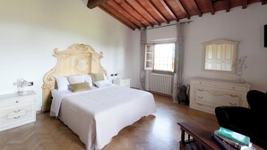 A bedroom with a large bed, wooden ceiling beams, a dresser, a nightstand, and a window with curtains.