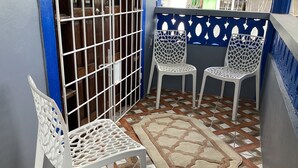A small, enclosed balcony with a tiled floor, two white plastic chairs, a patterned rug, and a barred door.