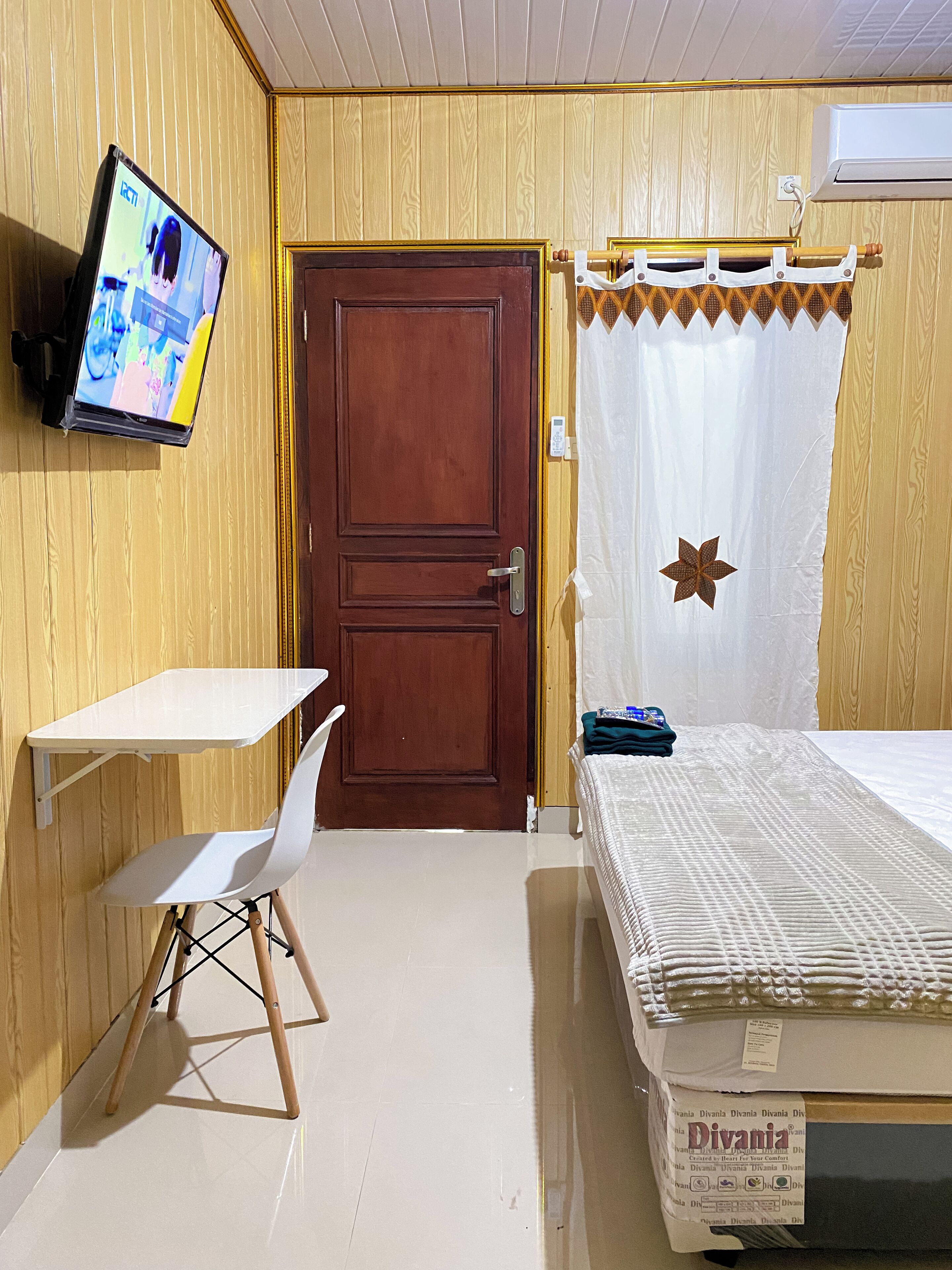A room with a wooden paneled wall, a white desk with a chair, a bed with a white duvet, and a television mounted on the wall.