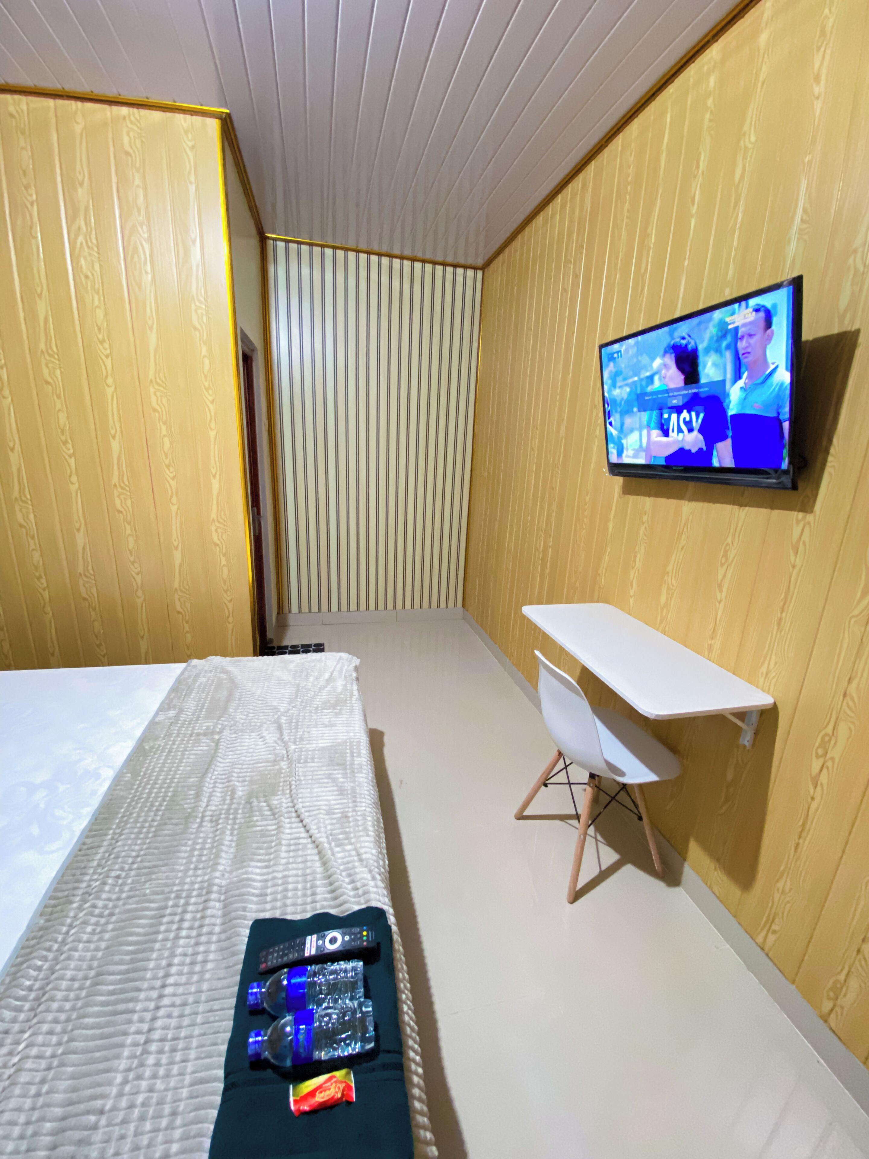 A hotel room with a bed, a desk, a TV mounted on the wall, and a window with blinds.