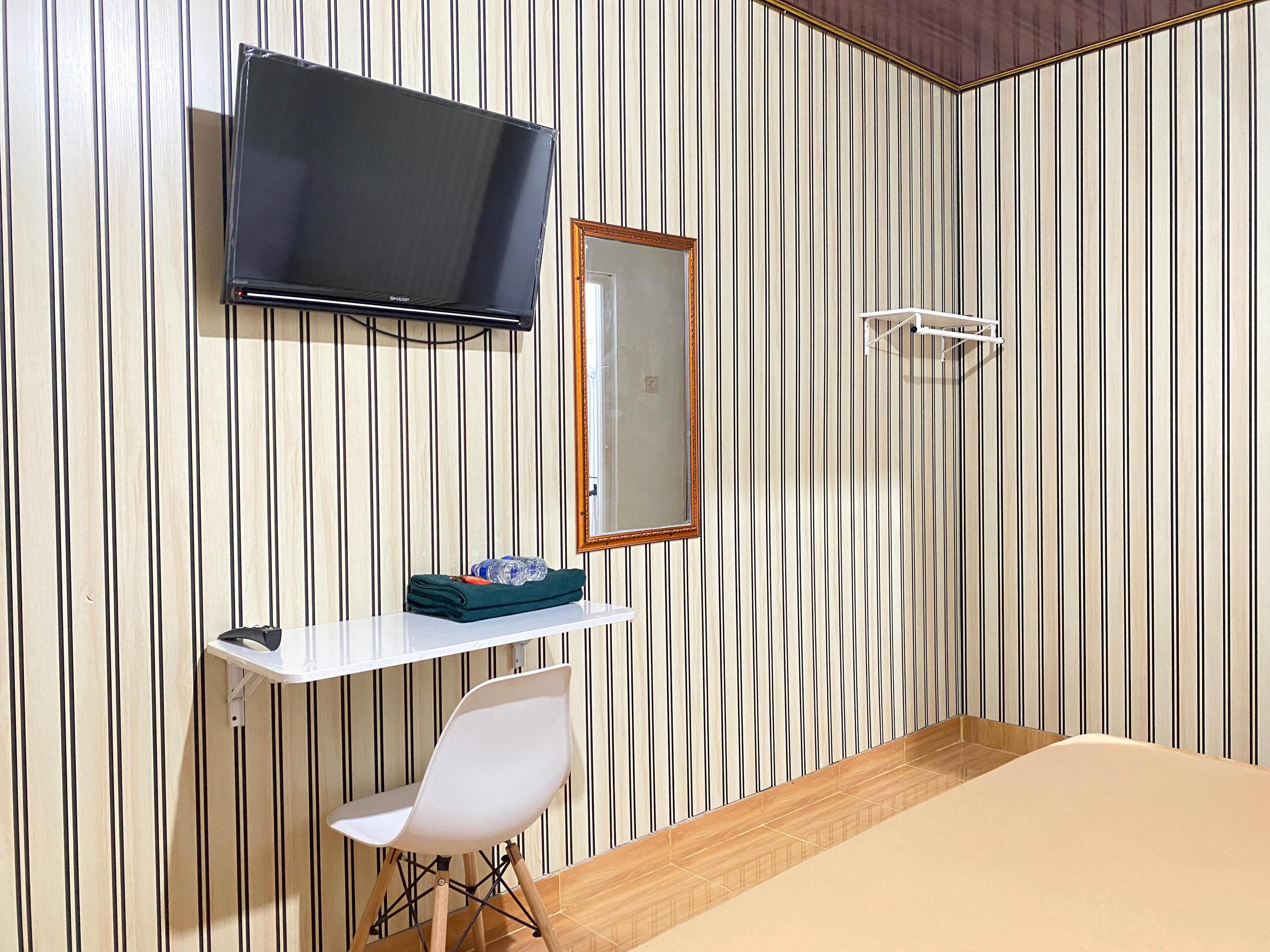 A hotel room with a striped wallpaper, a flat-screen TV mounted on the wall, a small white desk with a chair, and a bed with a wooden headboard.