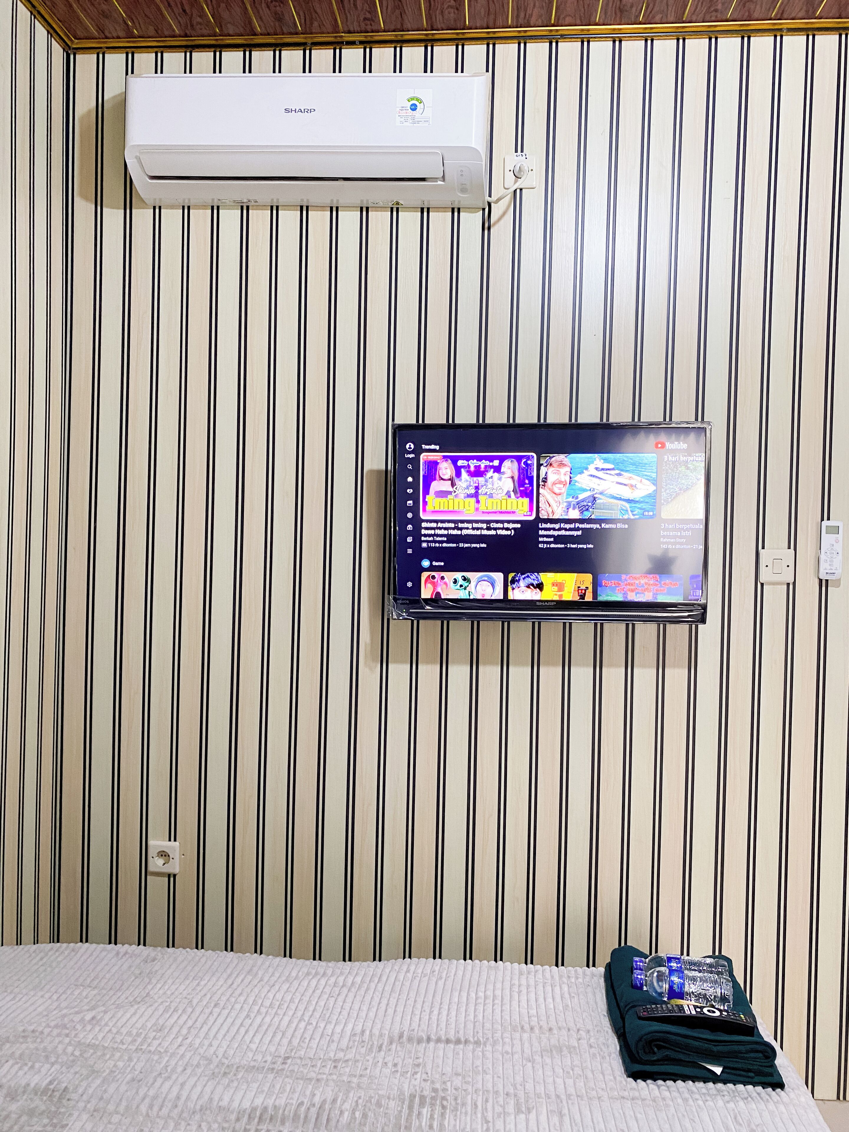 A room with a mounted air conditioner, a flat-screen TV displaying a YouTube channel, and a bed with a folded blanket and remote control.