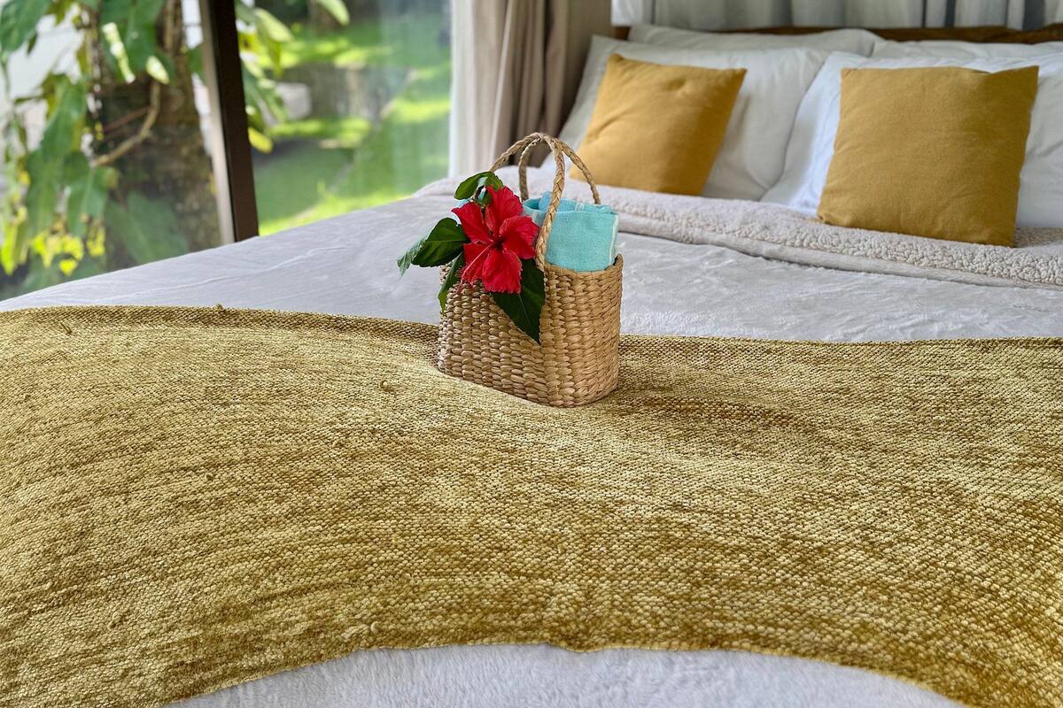 A bedroom with a bed, a woven basket with a red flower, and a view of greenery outside.