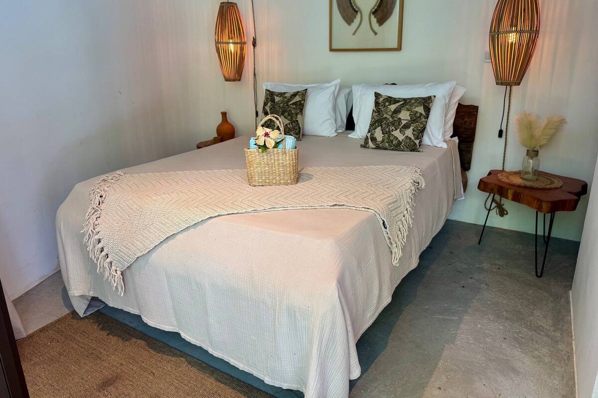 A neatly made bed with a white bedspread and pillows, a woven basket with flowers, and two wall-mounted lamps.