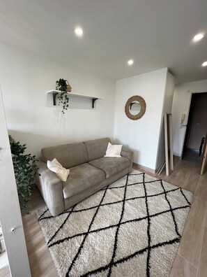 Een woonkamer met een beige bank, een geometrisch vloerkleed, een wandplank met een plant en een spiegel.
