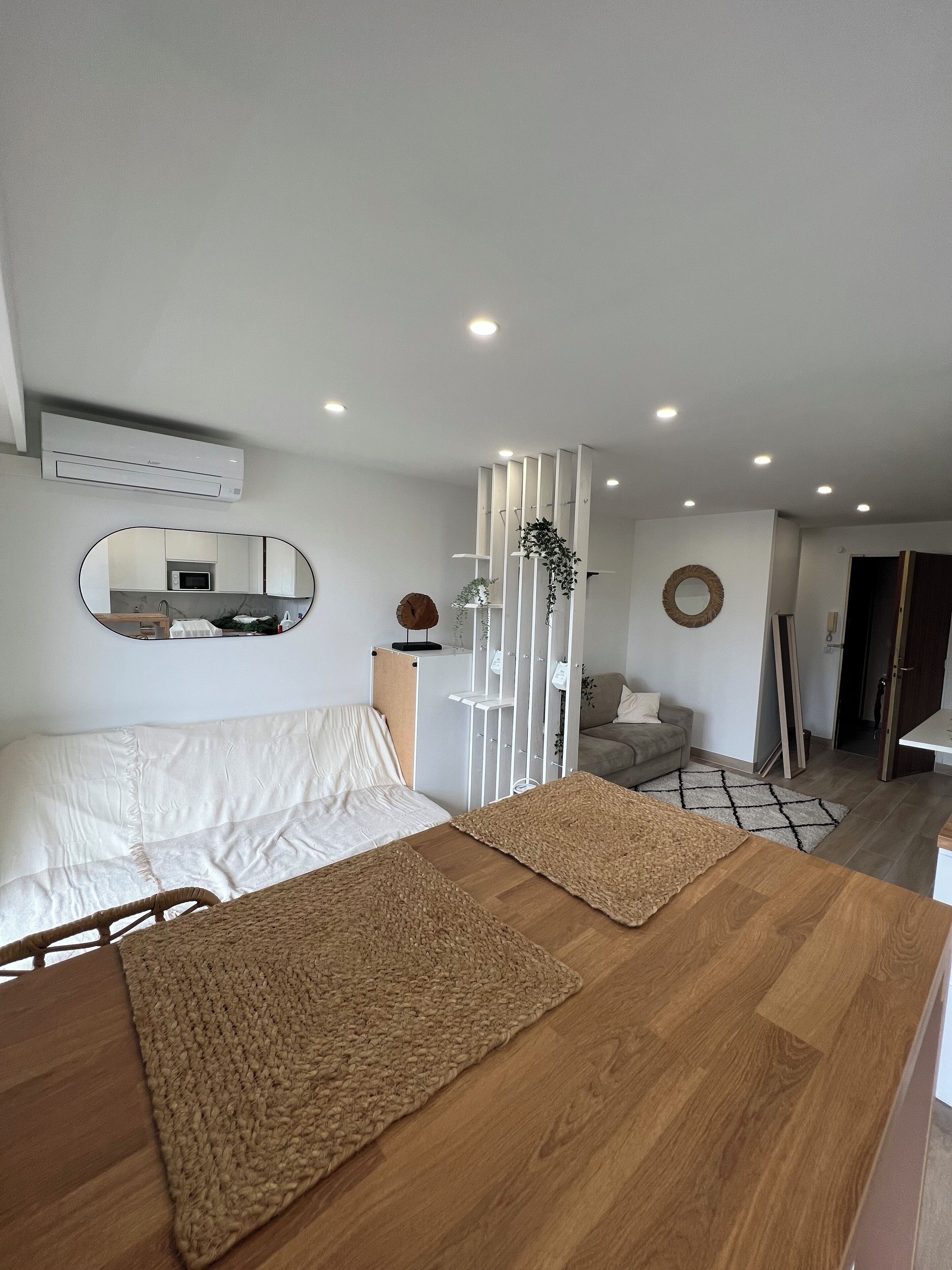 A modern bedroom with a large bed, a wooden bedside table, a mirror, and a view of a kitchen and living area.