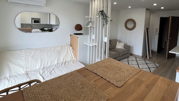Une chambre moderne avec un grand lit, une table de chevet en bois, un miroir et une vue sur la cuisine et le salon.