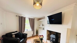 Un salon avec un canapé en cuir noir, une table basse, un téléviseur à écran plat fixé au-dessus d’une cheminée et une fenêtre avec des rideaux.