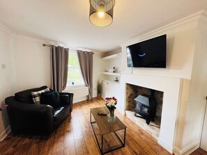 A living room with a black leather sofa, a coffee table, a flat-screen TV mounted above a fireplace, and a window with curtains.