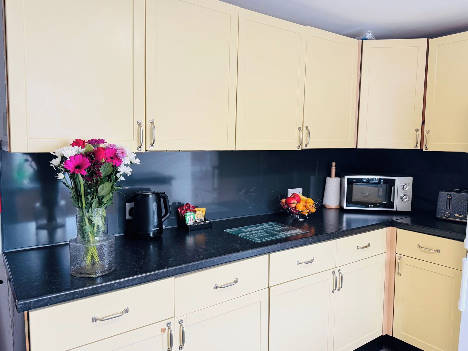 Eine Küche mit cremefarbenen Schränken, einer schwarzen Arbeitsplatte, einer Kaffeemaschine, einer Mikrowelle, einem Toaster und einer Vase mit Blumen.