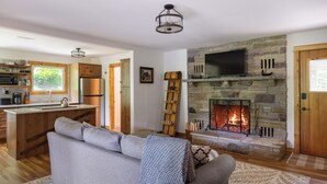A cozy living room with a fireplace, a sofa, and a kitchen area with wooden cabinets and a window. - Waterfront retreat with fireplace 3 miles from Whiteface & The North Pole (Wilmington)