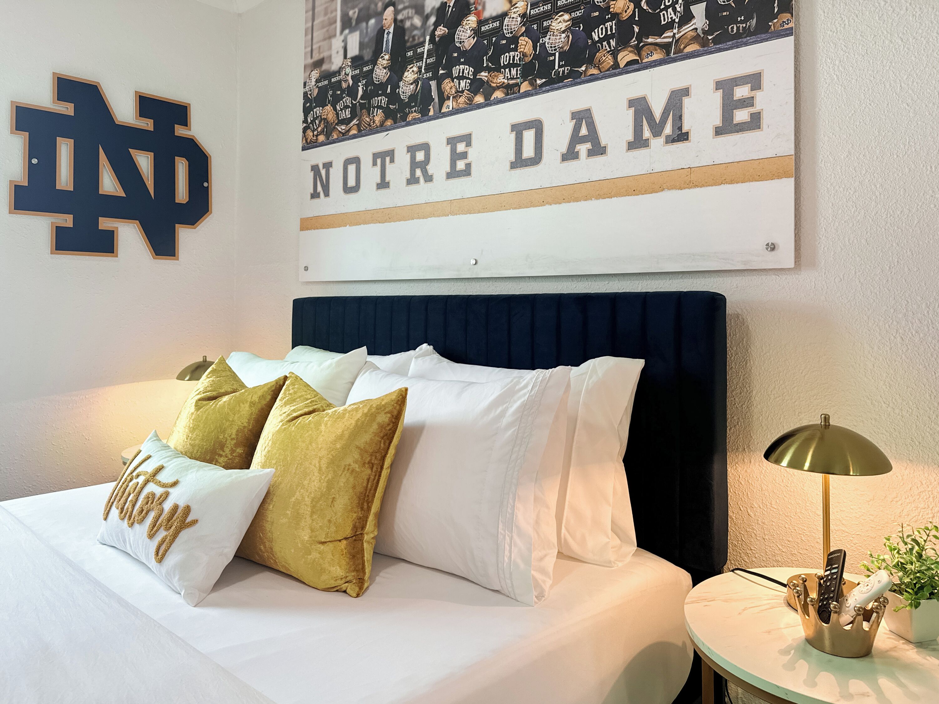 Een hotelkamer met een bed, een bord met Notre Dame erop en een lijst met foto’s van American footballspelers van Notre Dame.