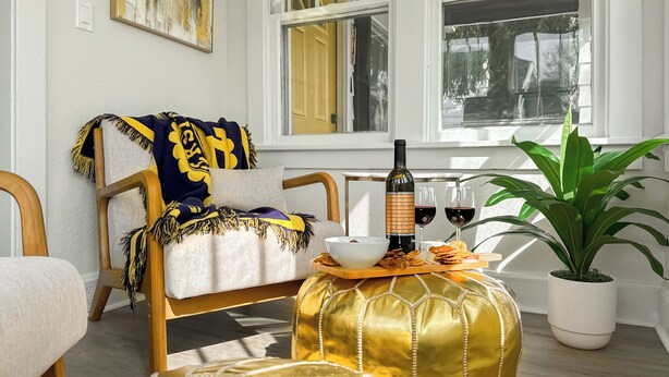 Un salón acogedor con un sillón cómodo, un otoman decorativo, una botella de vino, copas y una planta en maceta.