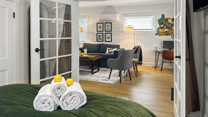 Una sala moderna con un sofá, una mesa de centro y un comedor con sillas. Se ve una cama con ropa de cama verde y toallas enrolladas.