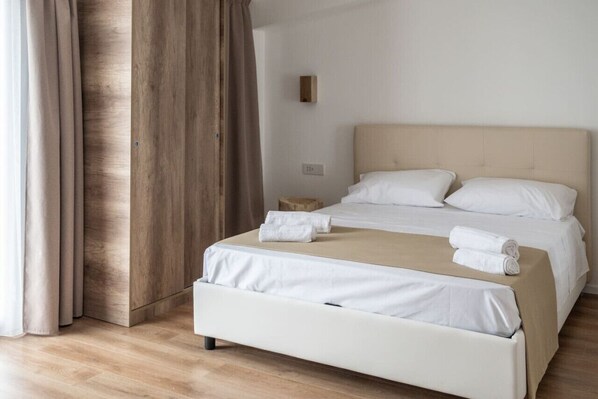 Una cama bien hecha con sábanas blancas y un cabecero color beige, una mesita de noche de madera y un interruptor de luz en la pared.